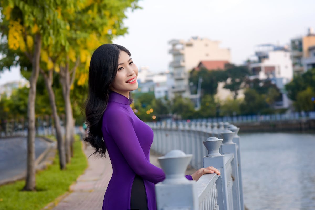 Smiling woman in a purple Ao Dai standing by a scenic waterfront on a sunny day.